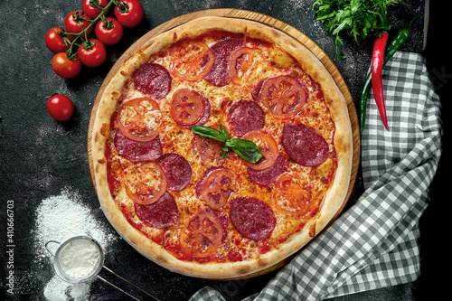 Oven-baked Italian pizza with red sauce, cheese, tomatoes and pepperoni salami in a composition with ingredients on a dark background. View from above.