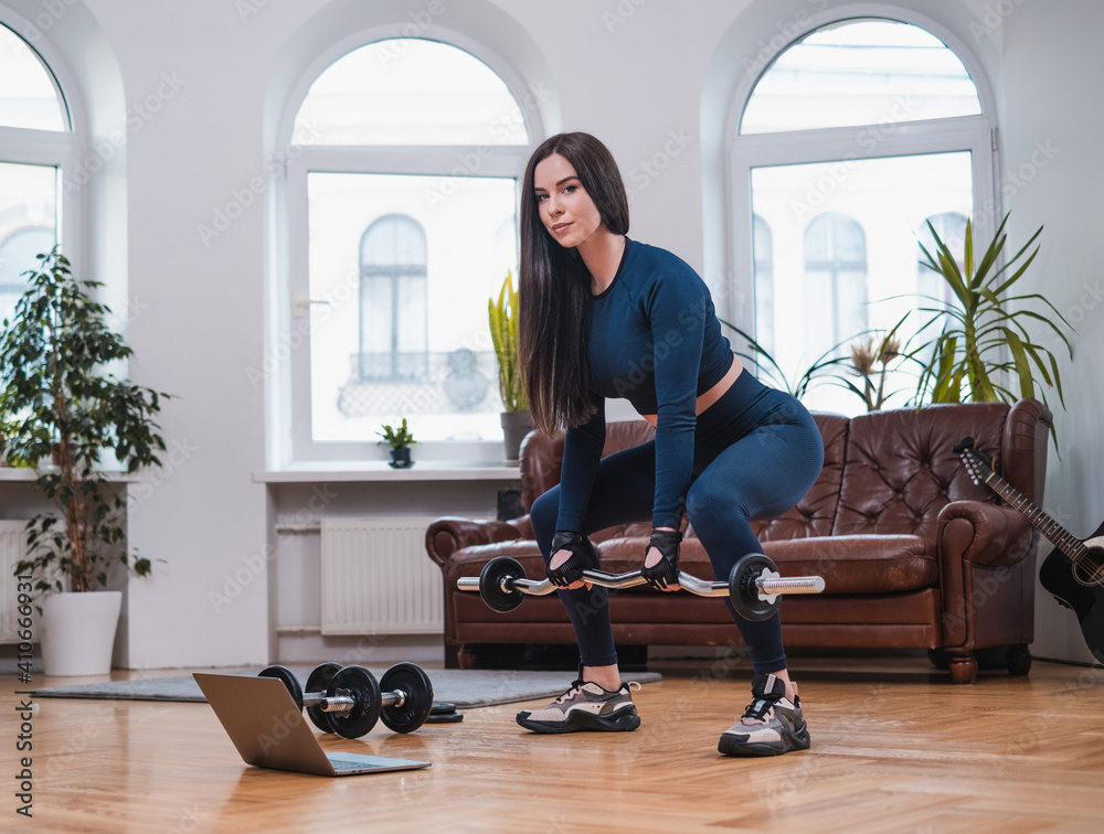 Strong girl with barbell and laptop works out in cosy and modern apartment with furniture. Strong girl with barbell and laptop works out in cosy and modern apartment with furniture.