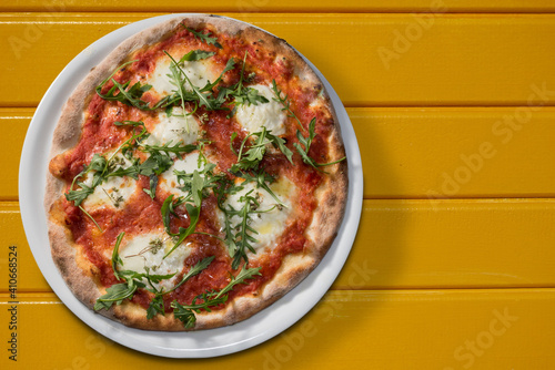 Pizza with tomato sauce, mozzarella and arugula or rocket in white plate on yellow boards, flat lay in top view with copy space