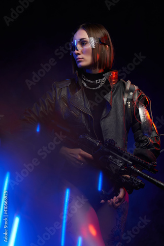 Tattooed female soldier with eyewear and modern hairstyle poses in dark background with lights. Glamour and at the same time dangerous woman with cybernetic shoulder.