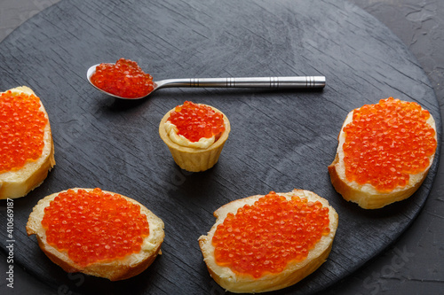 Canapes with butter and red caviar and a spoon with caviar on a black round board on a concrete background copy space