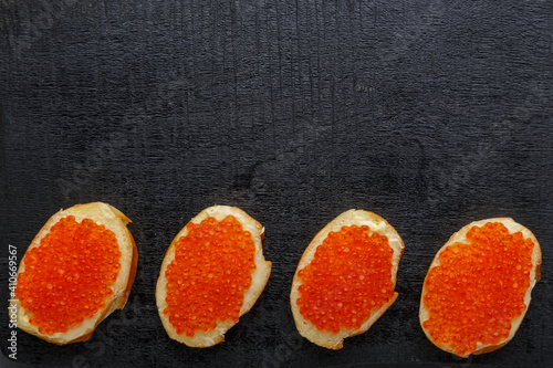 Bruschetta with butter and red caviar and a spoon with caviar on a black board in a row. copy space.