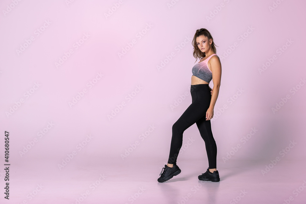 Young fit female trainer standing one foot on toes showing exercise, sporty woman ready for training with pink background