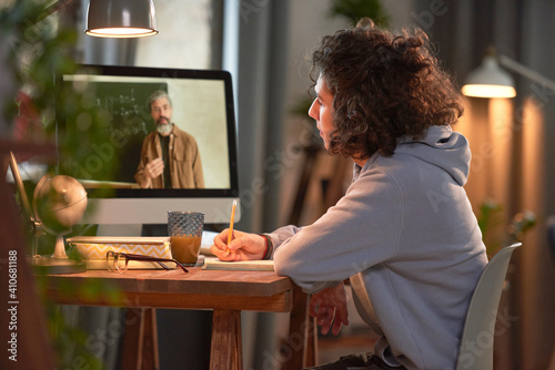 Student sitting at the table in front of computer monitor and listening to his teacher during online studying photo