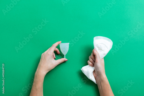 Female hands holding menstrual cup and crumpled sanitary pad over green background with copy space