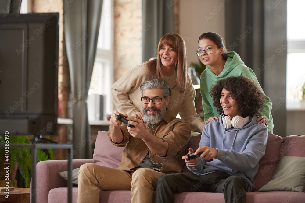 Father and son playing video game on sofa with daughter and mother supporting them in the background Father and son playing video game on sofa with daughter and mother supporting them in the background