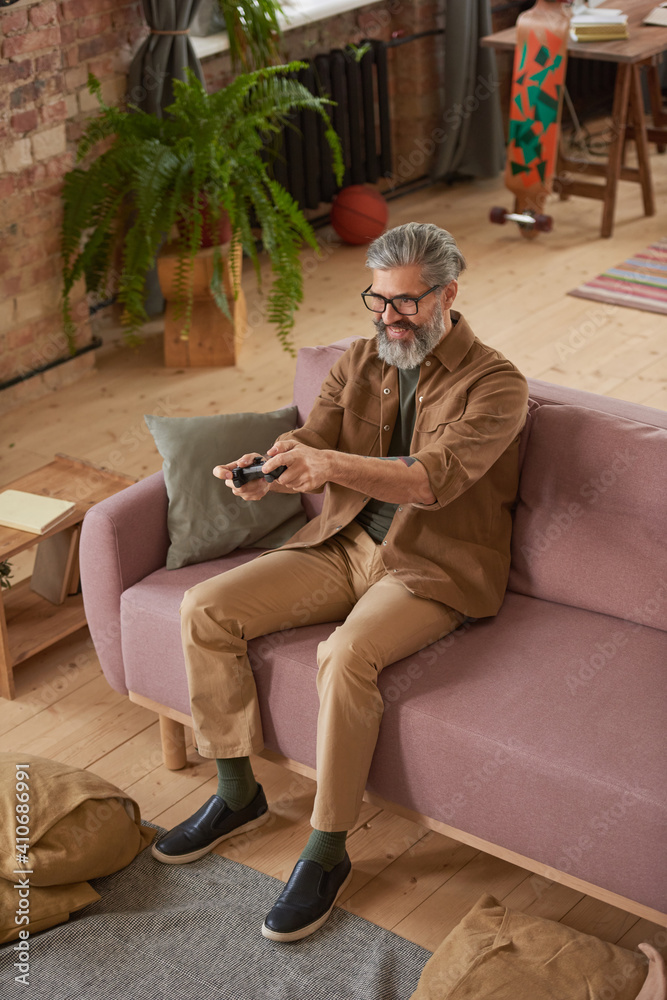 Mature bearded man sitting on sofa and playing online in video games in the living room at home