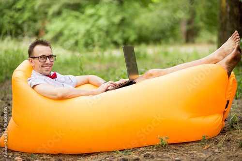 Young stylish freelancer man in glasses working at laptop sitting on Inflatable mattress in garden. Distance learning outdoor. Online education by Internet.