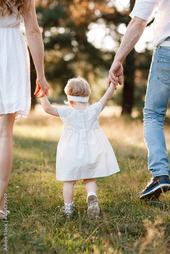 Fototapeta premium View on toddler. Back view of mother, father hold hands daughter enjoy nature and walk in the summer park. Young family spending time together on vacation, outdoors. Mother's, father's, baby's day.