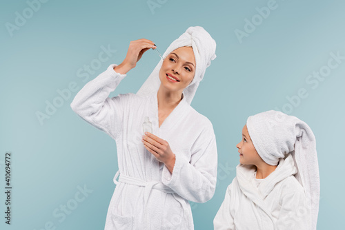happy mother in bathrobe holding bottle and applying serum near child isolated on blue