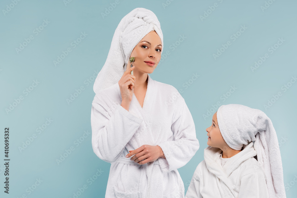 happy kid in bathrobe looking at mother using jade roller isolated on blue happy kid in bathrobe looking at mother using jade roller isolated on blue