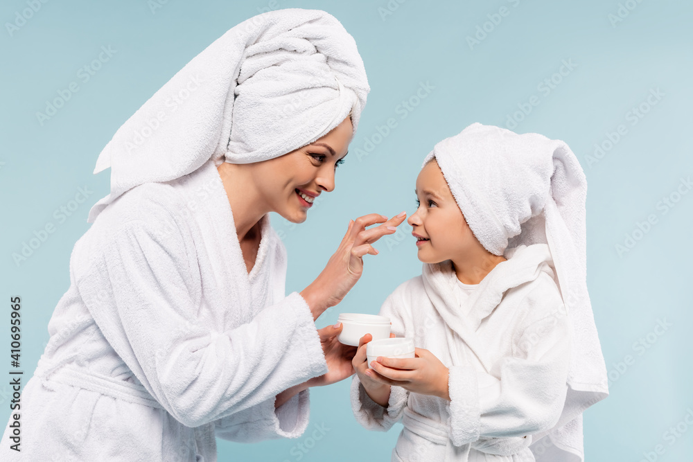 happy mother in bathrobe holding container and applying cosmetic cream on face of daughter isolated on blue