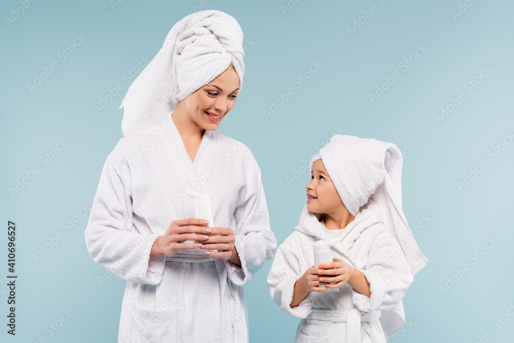 happy mother and child in bathrobes holding tubes with cosmetic cream isolated on blue happy mother and child in bathrobes holding tubes with cosmetic cream isolated on blue