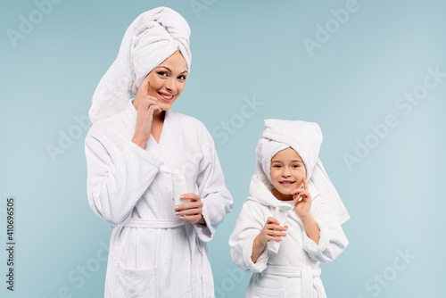happy mother and kid in bathrobes holding tubes and applying cosmetic cream on faces isolated on blue