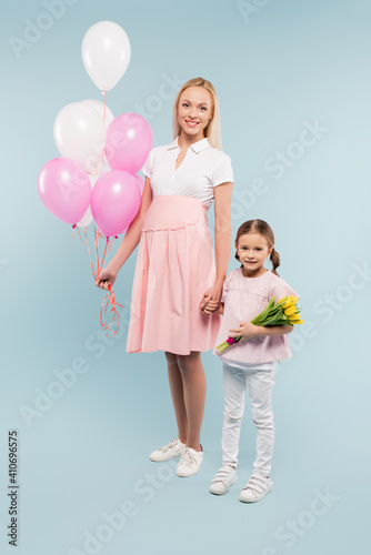 full length of happy pregnant woman holding balloons near daughter with tulips on blue