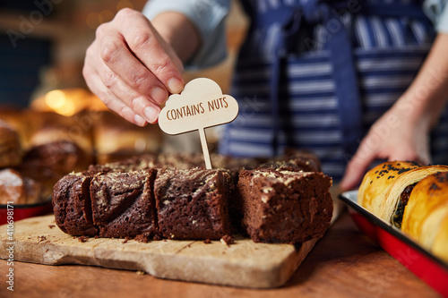 Sales Assistant In Bakery Putting Contains Nuts Label Into Stack Of Freshly Baked Baked Brownies photo