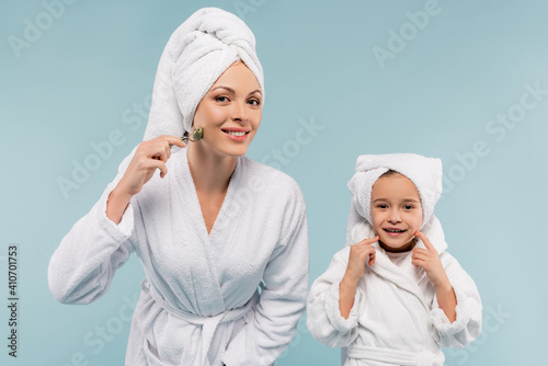 happy kid in bathrobe smiling near mother using jade roller isolated on blue