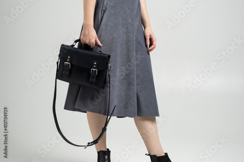 Small black leather bag in a woman's hand on a white background. Shoulder handbag. Woman in a white shirt and grey skirt and with a black handbag. Style, retro, fashion, vintage and elegance.