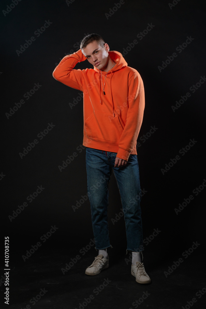 Young handsome man in jeans and orange hoodie posing while standing on a black background.
