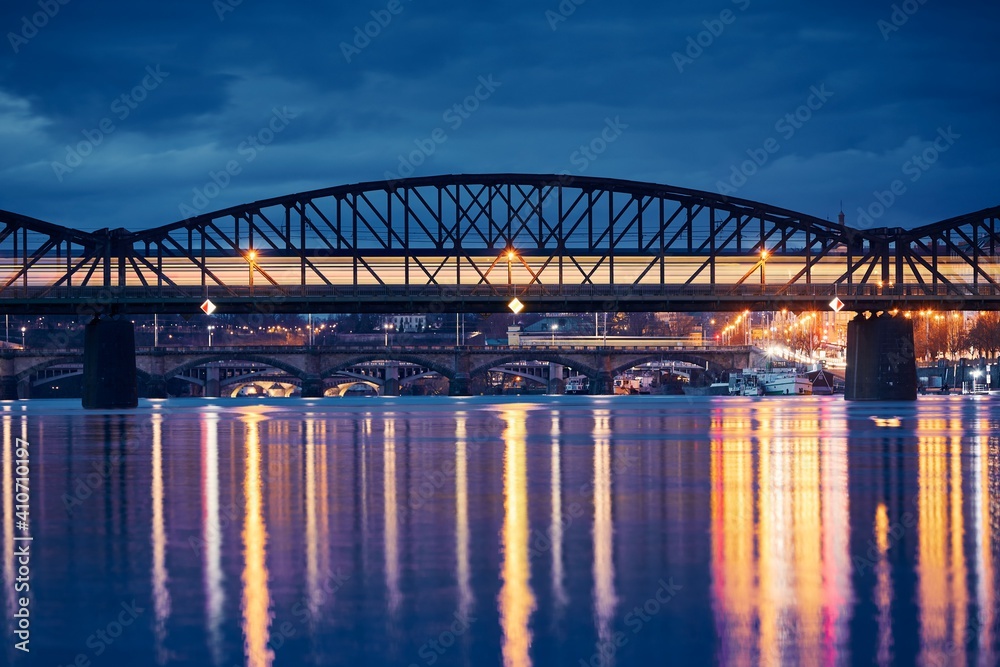 Fototapeta premium Passenger train passing over railway bridge against city. Cityscape of Prague at night, Czech Republic