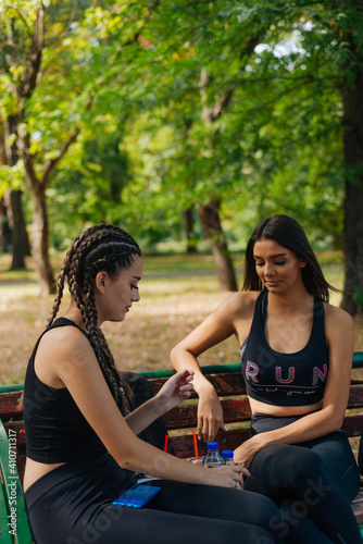Two girls sit in a park while taking a break from rollerskating drinking smoothie water and gossiping. Girls bonding concept.