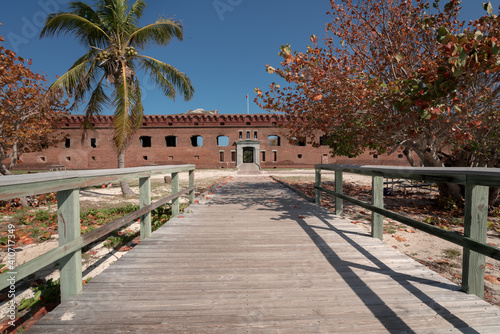 Gate of an old military fort with a wooden walk in the foreground. Big brick walls and green palm trees. Fort Jefferson.