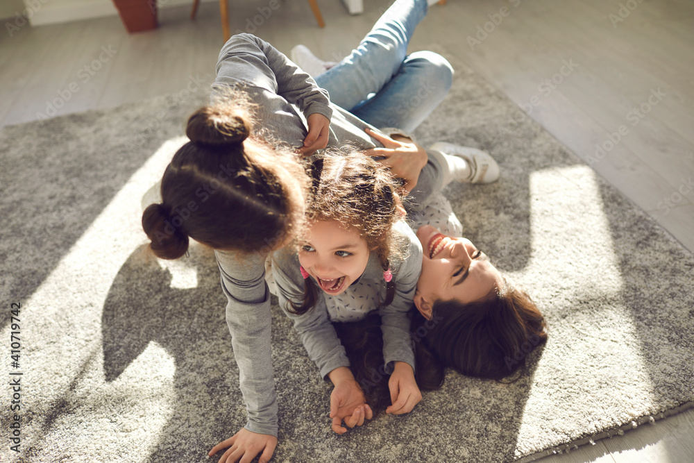 Happy family enjoying quality time at home. Mom and little children frolicking on carpet. Cheerful mother and funny carefree kids playing, laughing and having fun on floor rug in sunny living-room - obrazy, fototapety, plakaty 