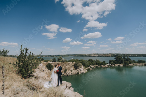 Wedding portrait of newlyweds in love in nature. Stylish groom in a blue suit and a beautiful bride in a long white lace dress embrace against the backdrop of rocks  cliffs  sky and river.