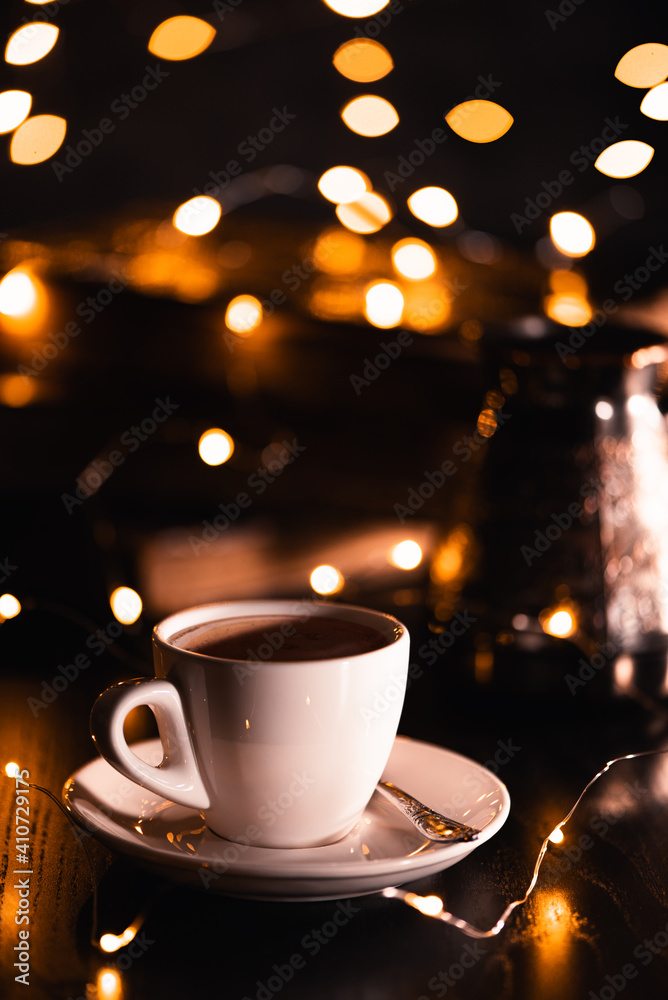 White cup of coffee on dark wooden background in yellow defocused lights of garland. Festive mood. Holiday concept.