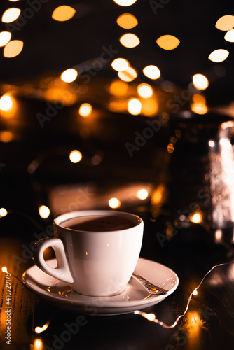 Obraz na płótnie White cup of coffee on dark wooden background in yellow defocused lights of garland. Festive mood. Holiday concept. w salonie