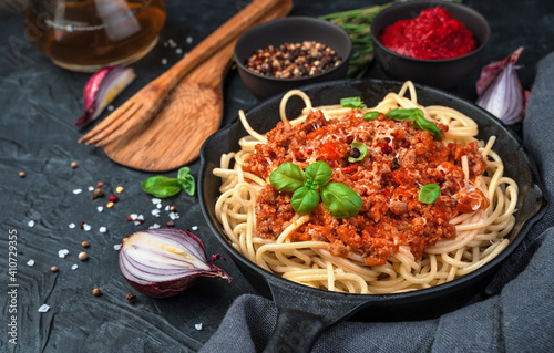 Spaghetti and sauce in a frying pan are sprinkled with cheese and fresh basil on a background of spices and onions on a black concrete background. Side view, horizontal.