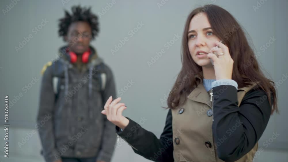 Portrait of irritated young Caucasian woman talking on the phone as blurred African American courier waiting at background. Dissatisfied client arguing with delivery service. Lifestyle. Portrait of irritated young Caucasian woman talking on the phone as blurred African American courier waiting at background. Dissatisfied client arguing with delivery service. Lifestyle.
