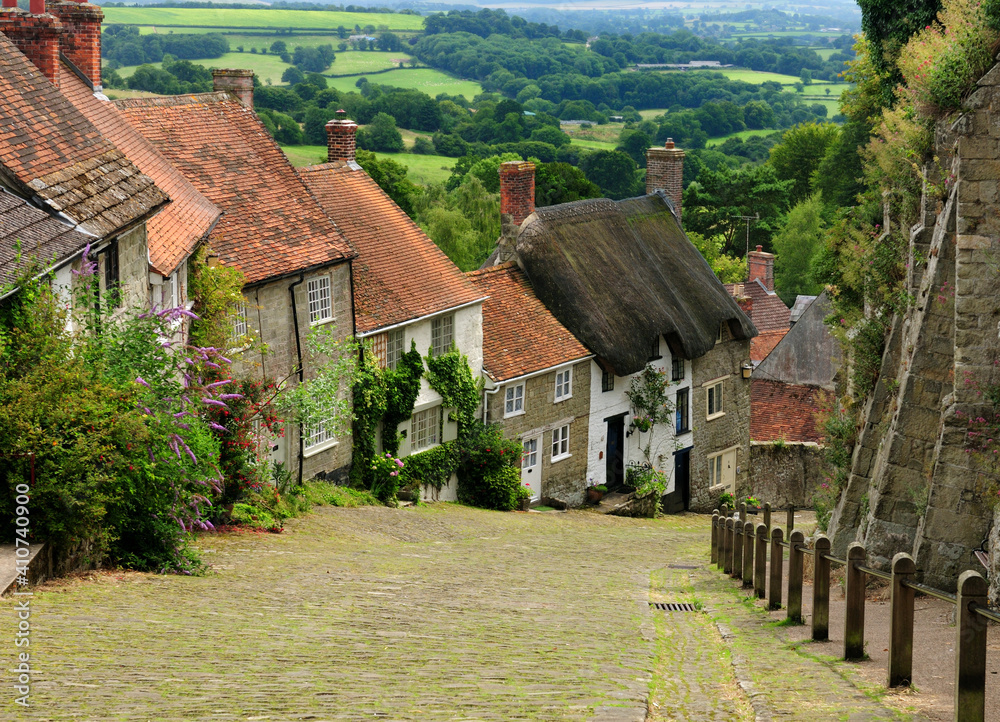 View To Old Limestone Houses On A Cobble Street At Gold Hill In Shaftesbury England With A Beautiful Rural Landscape In The Background On An Overcast Summer Day