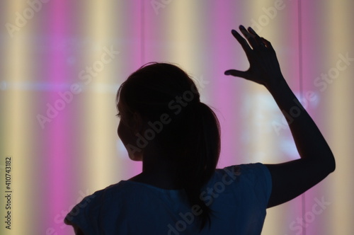 A young woman is raising her hand looking to the left. We see her from back and don't see her face. The background is colourful neon wall.
