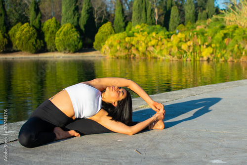Young attractive sportswoman practicing yoga, doing the exercise Revolved Head to Knee Forward, Parivrtta Janu Sirsasana pose, exercising, in the morning and a lake in the background.
