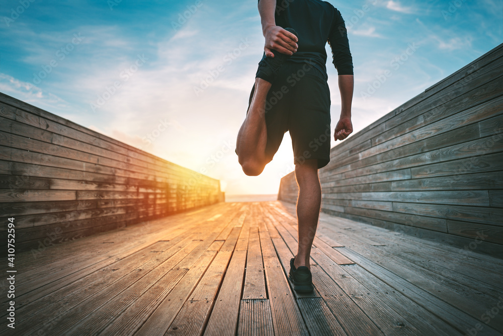 Young slim sporty man warming and stretching legs before run outdoors at sunset or sunrise. Athletic man in black sportswear doing fitness stretching exercises. Sport and healthy lifestyle. - obrazy, fototapety, plakaty 