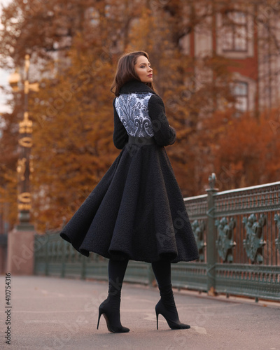 Elegant woman wearing black coat and walking city street on autumn or fall day against town park fence. Pretty girl with makeup and wavy brunette hair