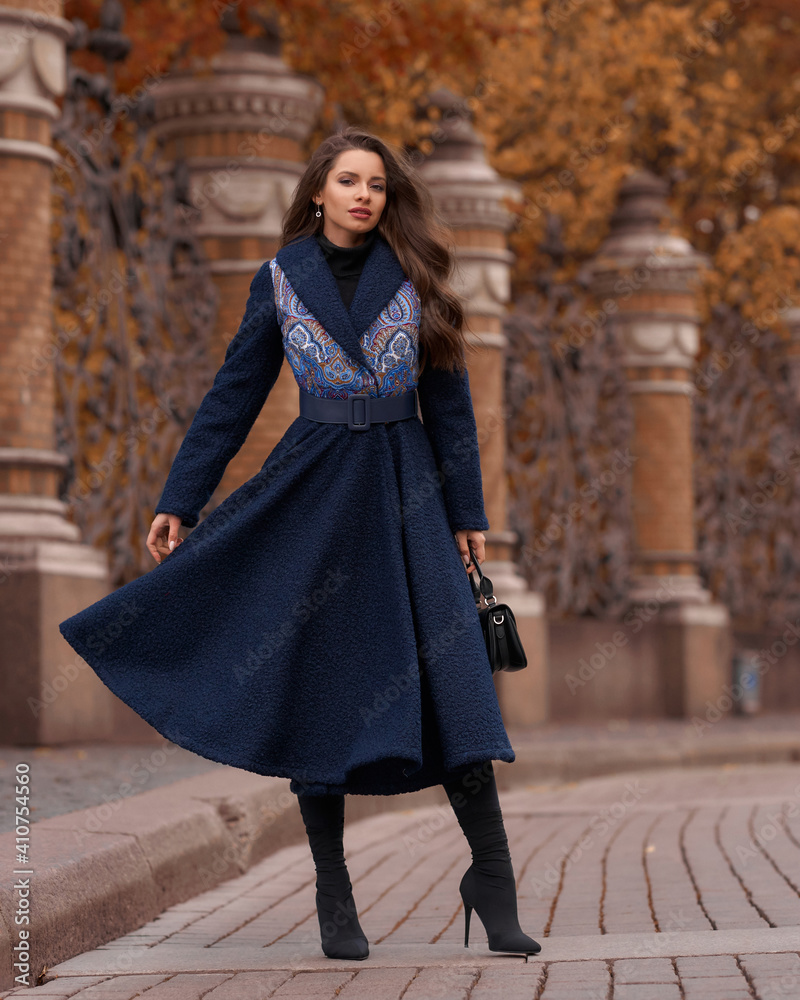 Elegant woman wearing blue coat and walking city street on autumn or fall day against town park fence. Pretty girl with makeup and wavy brunette hair