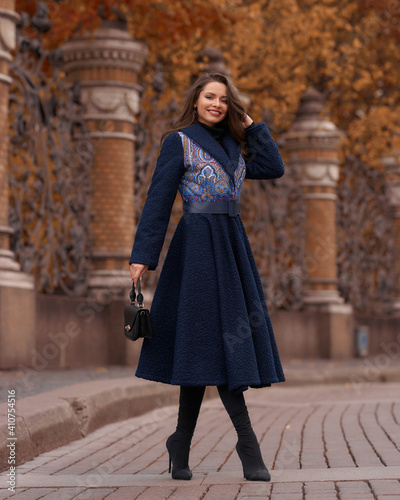 Elegant woman wearing blue coat and walking city street on autumn or fall day against town park fence. Pretty girl with makeup and wavy brunette hair