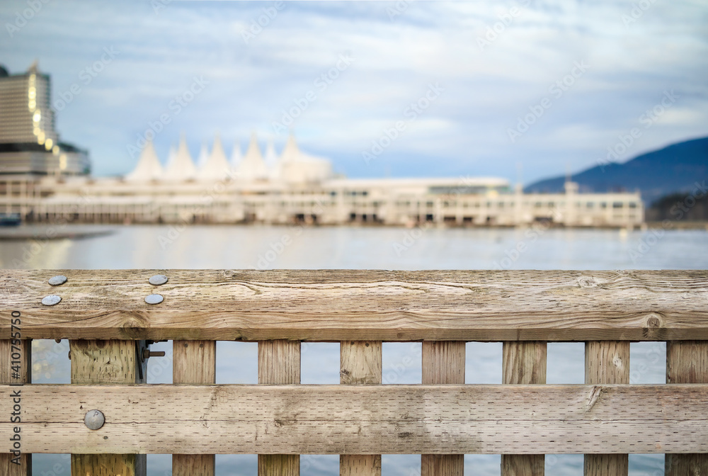Fototapeta premium Weathered wood railing with blurry waterfront buildings and ocean background and cloudy sky. Defocused and abstract Vancouver Harbour building or Canada Place. Selective focus.