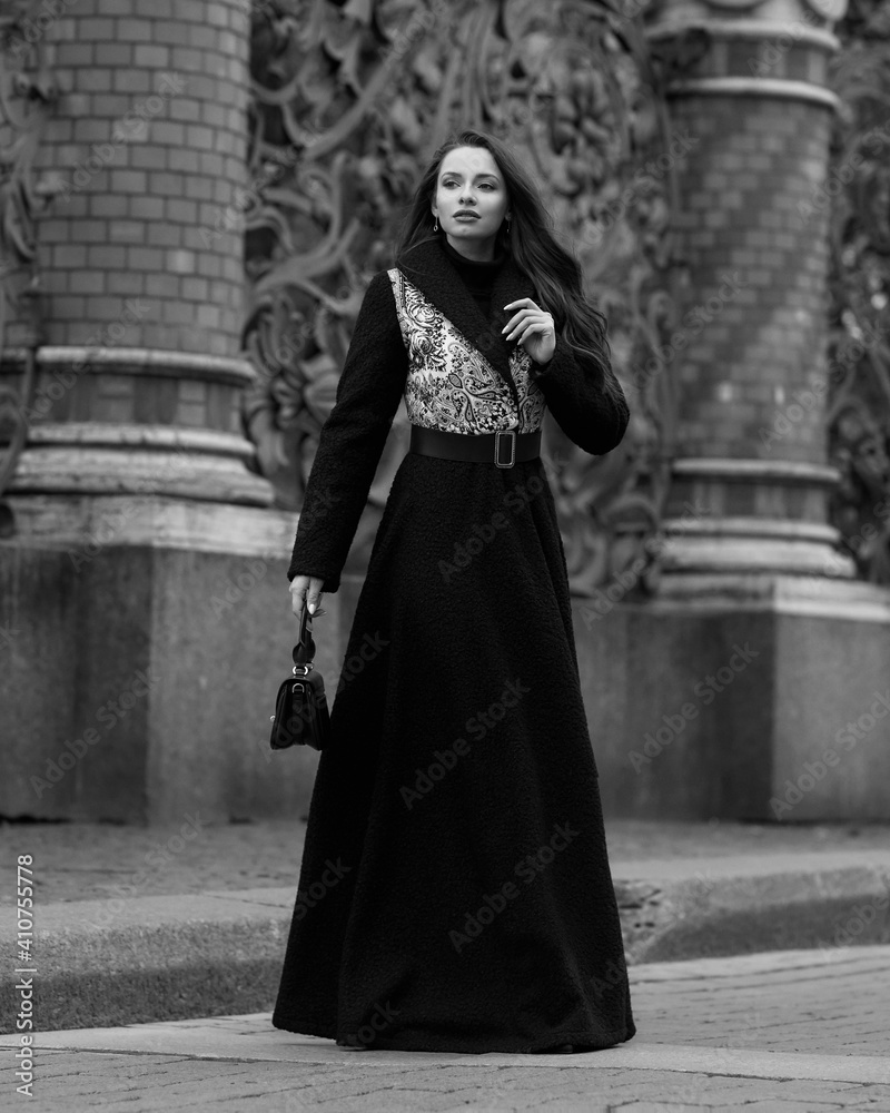 Elegant woman wearing black coat and walking city street on autumn or fall day against town park fence. Pretty girl with makeup and wavy brunette hair Elegant woman wearing black coat and walking city street on autumn or fall day against town park fence. Pretty girl with makeup and wavy brunette hair