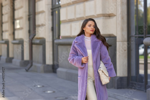 Elegant stylish woman with long brunette wavy hair wearing white trousers, pullover and shoes and purple fur coat walking city street on a sunny day