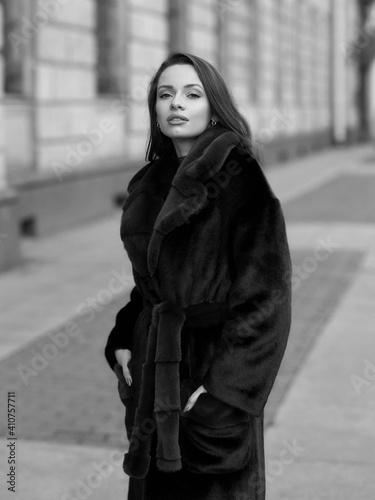 Elegant stylish woman with long brunette wavy hair wearing black trousers, pullover, shoes and fur coat walking city street on a sunny day © Dmitry Tsvetkov