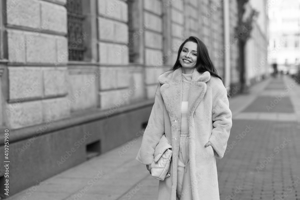 Naklejka premium Elegant stylish woman with long brunette wavy hair wearing white trousers, pullover and shoes and pink fur coat walking city street on a sunny day