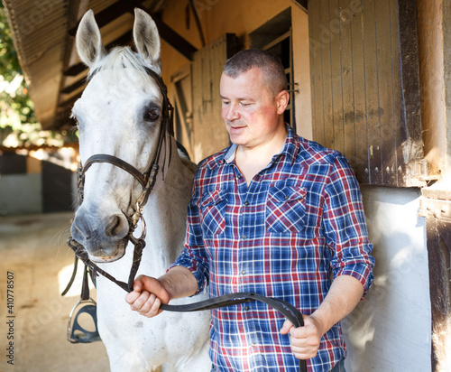 Positive male farmer standing with white horse at stable outdoor