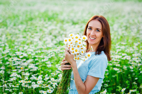 A young smiling brunette in a blue dress holds a bouquet of daisies in her hands and sits on a chamomile field. He looks at the camera. Copy space.