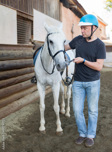 Mature smiling man farmer in helmet standing with white horse at stable outdoor