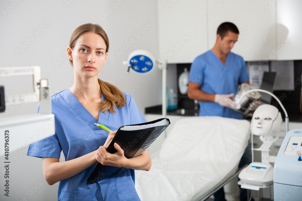 Obraz premium Young polite female cosmetologist meeting patient in office of aesthetic medicine, filling out medical form .