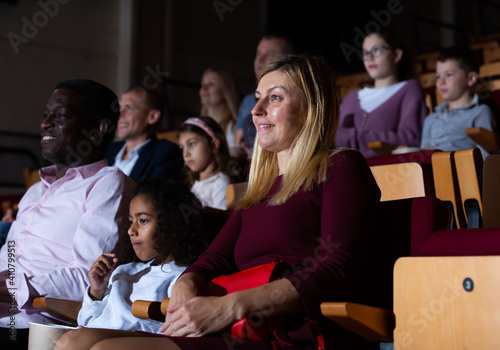 Friendly biracial family spending time together in cinema. Selective focus of young european woman sitting with cute preteen girl and aframerican man, absorbedly watching movie