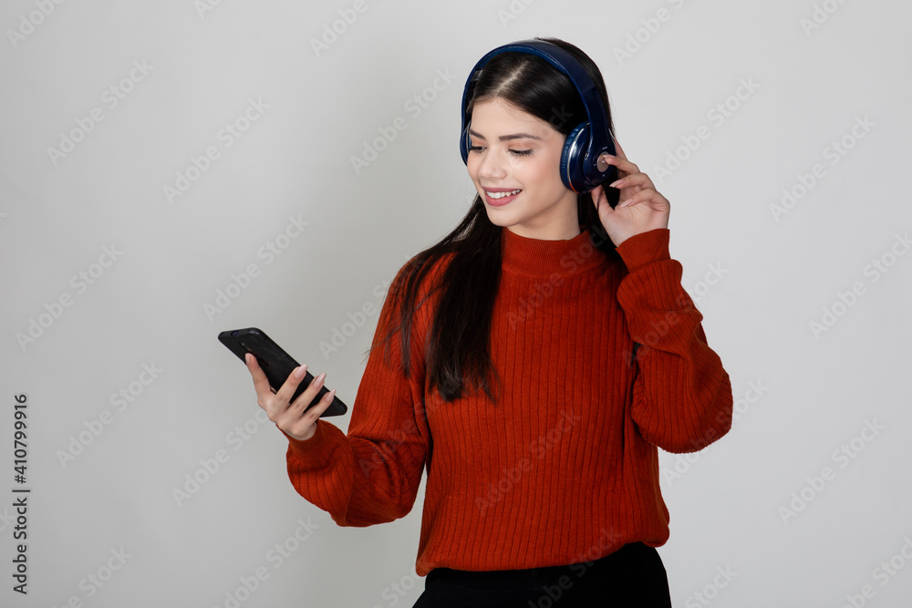 Pretty young girl holding her mobile and wearing headphone and listening to music or songs with a smile on her face, standing against white background. Pretty young girl holding her mobile and wearing headphone and listening to music or songs with a smile on her face, standing against white background.